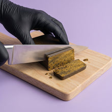 Découpe au couteau de chef d'un bloc de résine CBD Static Hash sur une planche en bois, montrant sa texture malléable