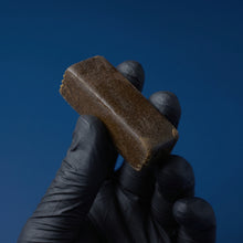 Main gantée de noir tenant un gros bloc rectangulaire de résine 3x filtré Maison Sativa, montrant sa texture externe brune, dense et brillante.