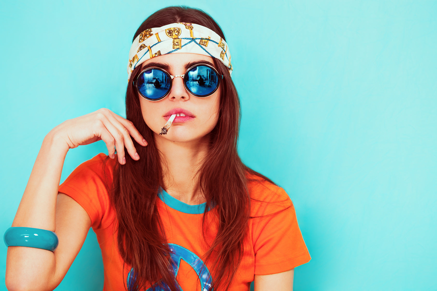 jeune femme habillée dans une couleur orange fluo comme dans les années 60 fumant un joint sur fond turquoise. 